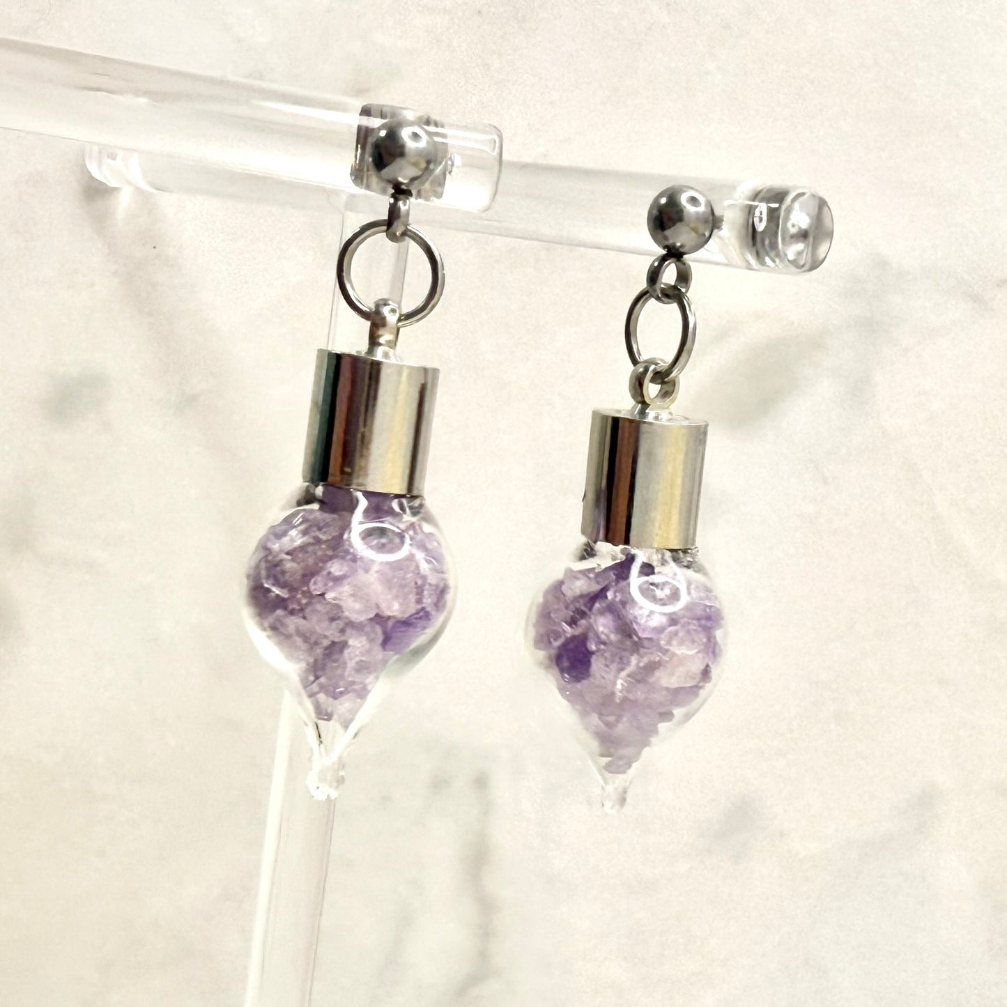 Pair of stud earrings with glass vial droppers filled with shards of natural amethyst hanging from a clear display hanger