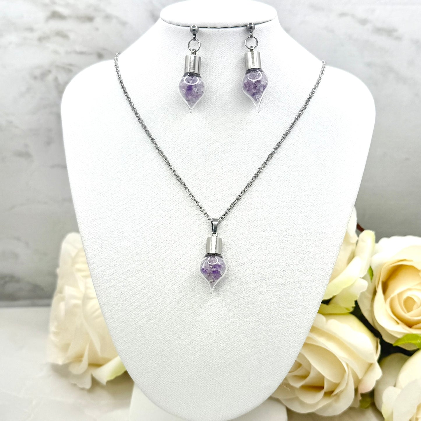 Jewelry set with glass vial filled with shards or natural amethyst including necklace and earrings on a mannequin bust with flowers in the background