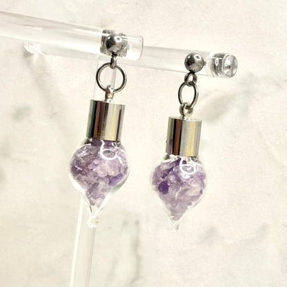 Pair of stud earrings with glass vial droppers filled with shards of natural amethyst hanging from a clear display hanger
