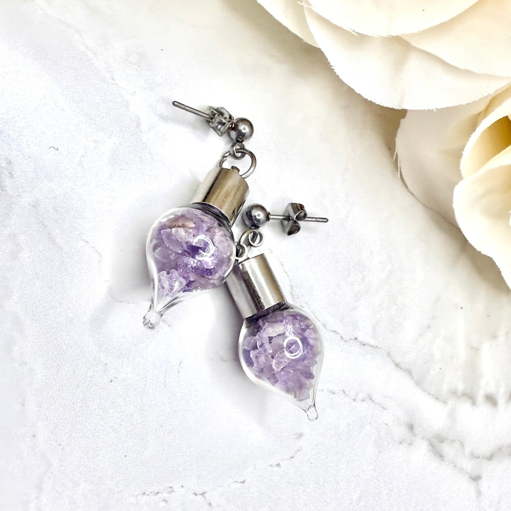 close up of pair of stud earrings with glass vials filled with shards of natural amethyst shown lying on their side with flowers in the background