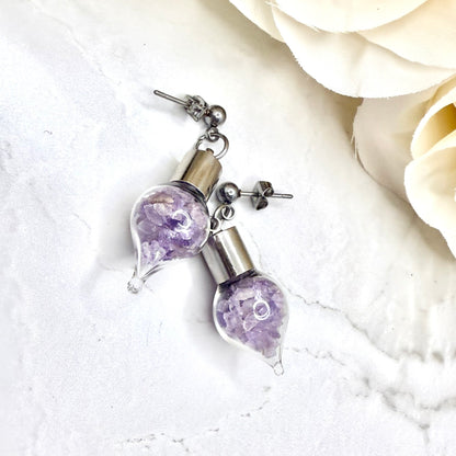 close up of pair of stud earrings with glass vials filled with shards of natural amethyst shown lying on their side with flowers in the background