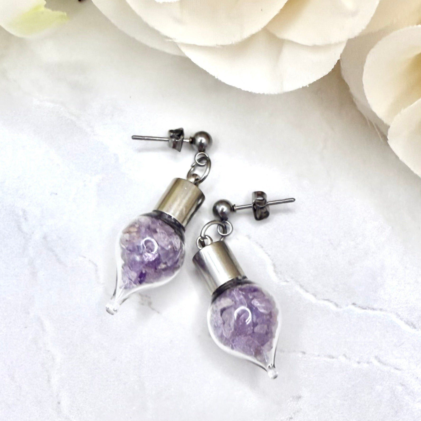 Pair of stud earrings with glass vials filled with shads of raw amethyst shown lying on a worktop with flowers in the background