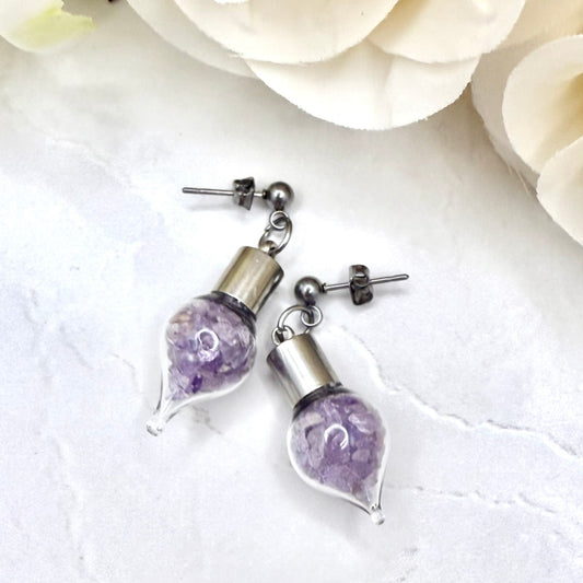 Pair of stud earrings with glass vials filled with shads of raw amethyst shown lying on a worktop with flowers in the background